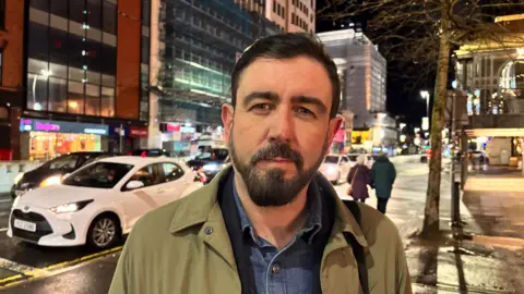 Paul Doherty. He has short dark hair and beard, wearing a khaki jacket with denim shirt underneath. He is standing on a street footpath with buildings lit up in the background and cars driving.