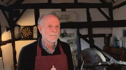 A man with grey hair and a grey beard is wearing a checked shirt, with a black jumper and a maroon apron. He is inside an historic room that is black and white timber framed. Behind him on the wall is a darts board.