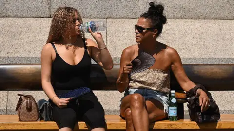 Two women are seen sat on a bench in summer clothing, one is drinking from a bottle of water and holding a fan on her lap, the other is wearing sunglasses and is fanning herself with a fan 