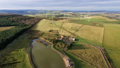 Huntly Development Trust A farm space with a large pond in the middle. There are wind turbines in the background with the rest of the space covered in grass. 