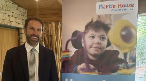 Sir Gareth Southgate stands smiling at the camera, wearing a dark suit with a white shirt and patterned tie. He is standing next to a banner which reads 'Martin House Children's Hospice' with a photo of a young child in a wheelchair underneath.