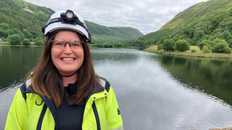 Sarah South wearing a hi-vis jacket and a white helmet and safety goggles, looking at the camera at the reservoir 