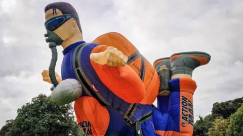 MJ Ballooning A large orange and purple Action Man hot air balloon is inflated in a grassy area surrounded by spectators and trees.