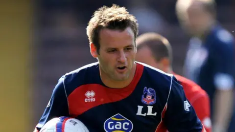 University of Chichester A man with short brown hair is running towards the camera while holding a football. He has a navy blue sports kit on with a red stripe