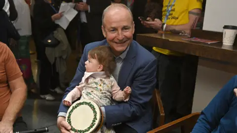 PA Media Micheál Martin is sitting with a toddler and a drum. He's indoors. People are in the background. 