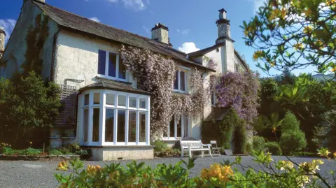 Rydal Mount is a white, two-storey house. Part of it is covered by purple shrubs and green fern growing along the entrance. A white bench and chair are outside the house, which is surrounded by trees.