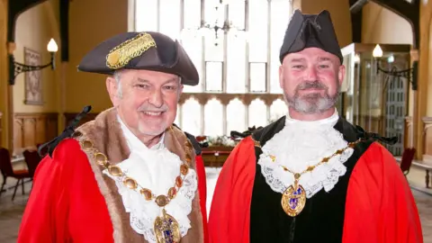 King's Lynn and West Norfolk Borough Council We can see two men dressed in mayoral outfits. They are both wearing red robes, tricorn hats and gold chains