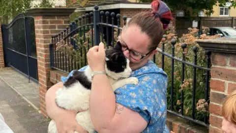 Lauren Mundy A woman in a blue flowery dress cuddling a black and white cat and burying her face in his fur