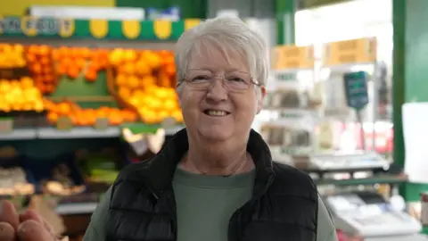 Barbara Farmer has short grey hair and glasses. She is wearing a green top and a black gillet and standing in a  greengrocers. 