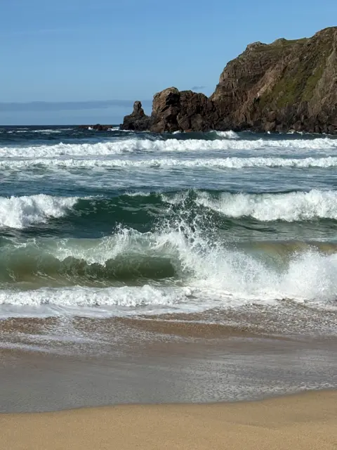 Wendy Greenstreet A sandy beach is bordered by churning waves rolling in from the sea. In the foreground, a breaking wave creates white spray and foam as it reaches the shoreline. Beyond the surf, multiple bands of waves stretch across the water towards a rugged rocky headland