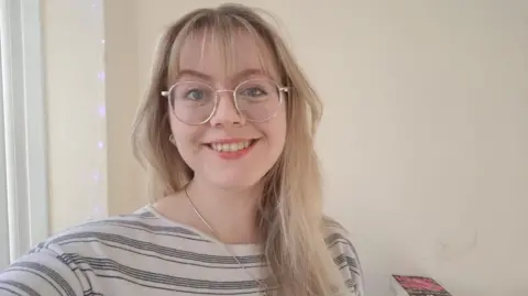 Catherine smiles at the camera, with long blonde hair, glasses, and a white top with black stripes. In the background are fairy lights and a pile of books.