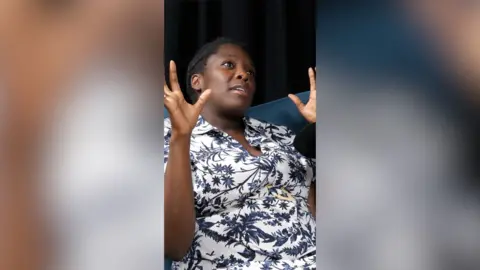 A woman with short, dark hair wears a floral blouse and talks to someone off-camera