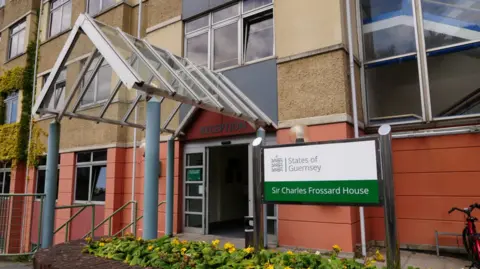 BBC Sir Charles Frossard House exterior, St Peter Port, Guernsey. A green and white sign is outside of the building. The sign says States of Guernsey Sir Charles Frossard House. The building is beige and red. A glass porch leads to the entrance of the reception. 
