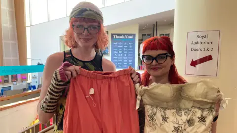 Alex Pope/BBC Two women holding up clothes. The young woman to the left has short pink hair, glasses and a bandana on her head. She is holding up pantaloons. The woman next to her has on glasses, long bright red hair and is holding up a vintage cream dress. 