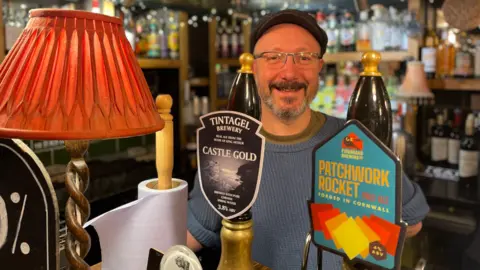 BBC Moth and Moon landlord Matt Reay stands behind the pub's bar. A lamp with an orange lampshade is on the bar. Two pumps with Castle Gold and Patchwork Rocket beers are visible. Mr Reay is wearing a blue jumper and a flatcap.