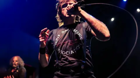 Dr David Gange Napalm Death's Barney Greenway sings into a microphone. He is wearing a Hunt Saboteurs Association tee-shirt and braces.