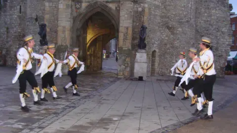 King John's Morris Men Six Morris men from King John's Morris Men wearing top hats white shirts, black trousers and white socks pulled up to the knee. They have yellow and black tassels and accessories. They are dancing in front of the bargate in Southampton at dawn