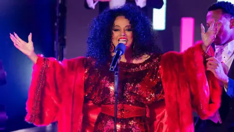 Getty Images A woman in a red dress and coat. She is singing on stage.