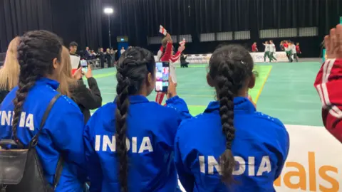 Girls from the Indian team watch a game with their backs to the camera. They have long dark hair which is tied back. Rival team members can be seen in the background.