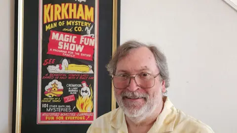 Supplied A man with a yellow shirt and glasses stands next to a post which says "Kirkham Man of Mystery and Magic Fun Show".
