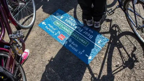 Getty Images Cyclists stand next to a sign painted on the floor marking the Two Tunnels Greenway cycle path. It shows directions to Midford, Radstock, Bath and Bristol. 