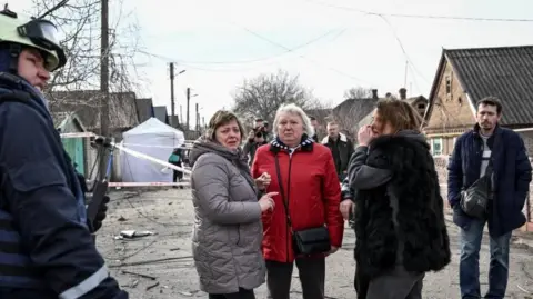 Three women pictured at the scene of the drone strike looking visibly upset. An emergency responder can be seen to their left and another man can be seen on the right. 
