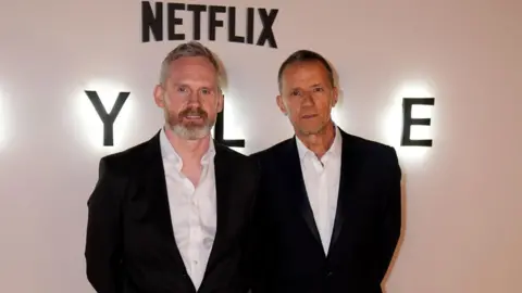 PA Media Two men in dark suits and white shirts standing in front of a white wall. Netflix is written on the wall above them.