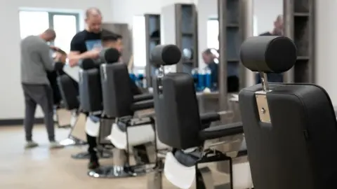 PA Media Row of folding seats with headrests and mirrors opposite. Two people stand up and cut their hair "customers" whose faces are blurred.