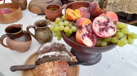 Militias of Anglia A display of food on a white tablecloth. A dark wood fruit bowl displays green grapes, pink pomegranates with red seeds, and dark pink peaches. There is slices bread on a wooden plate, and ceramic milk jugs