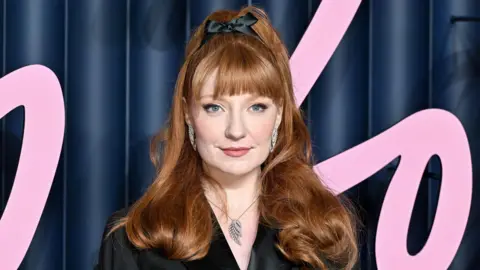 Getty Images Nicola Roberts wears a black top with a silver wing necklace and has her red hair tied up with a black bow, and stands in front of a dark blue background with pink swirls 