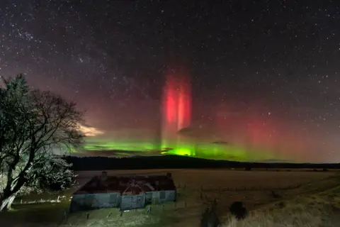 Alan Rankin Red and green coloured northern lights in a starry sky with an empty field and small stone farm building on the left with a large tree in the front of the house