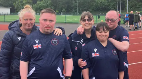 Grace Wood/BBC Two able bodies women - one blonde and on with brown hair - stand on a athletics track with two men and one woman who have Down Syndrome. They are wearing dark blue Team GB kit