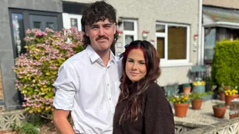Supplied Justyce and David standing in front of a row of houses with a pink flower bush behind them.