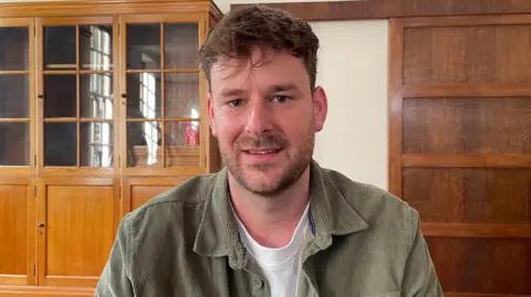 Ben Schofield/BBC Jack Cooper, looking straight to camera and smiling a little. To the left of the picture is a large empty, glass-fronted bookcase and on the right is a wooden panel. Jack has brown hair and is wearing an unbuttoned sage green corduroy shirt over a white t-shirt.