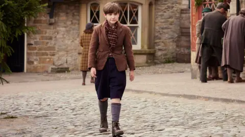 Playground/Channel 5 A photograph of Owen on set, wearing a brown jacket and scarf, knee-length trousers and long socks. He walks across a pebbled square, a sign that reads "Drovers Arms" can be seen on the wall in the background, partially obscured by a pine tree on the left. Three people are looking at produce on the right.