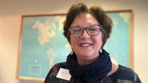 Emma Baugh/BBC Penny Heath has short brown hair and is smiling at the camera. She is wearing spectacles and a patterned top and blue scarf