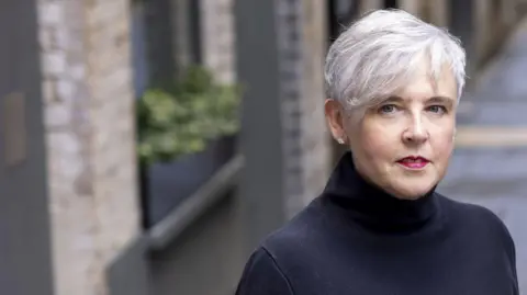Ruth Sparkes A woman with short, white hair and blue eyes is wearing diamond stud earrings and a black polo neck top. A blurred background behind her shows a wall and a window box with plants in it.