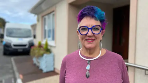 Mandy Le Bachelet, manager of Guernsey Voluntary Service, smiles while standing outside one of the charity's buildings. She has pink, purple and blue coloured hair and blue-rimmed glasses. She has grey dangly earrings and a large necklace on. She is wearing a pink jumper. A white minibus is in the background.