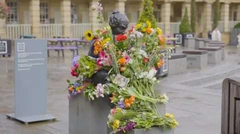 Calderdale Council Flowers on a grey statue of a woman. Behind here are benches on a city square.