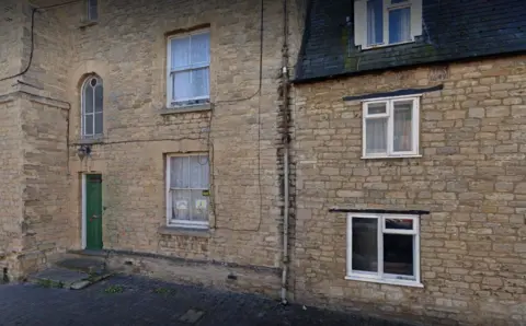 Google The side of a large brick building with windows on the ground and first and second floors. There is a green door with steps up to it and a path in front.