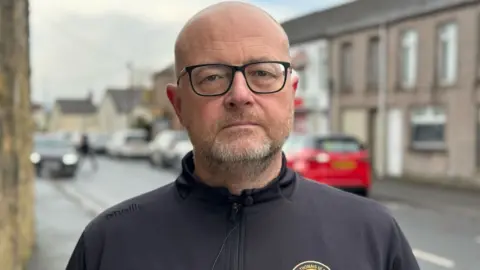 A man with a grey beard,dark rimmed glasses and a zip up top 