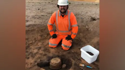 Oxford Cotswold Archaeology Will, an archaeologist in a hi-vis orange suit and wearing a white hard hat, kneeling in front of a just-excavated Bronze Age collared urn found on Sizewell C excavation site in Suffolk and smiling.  On the right is white plastic box with earth or other finds in it and a paint brush. 