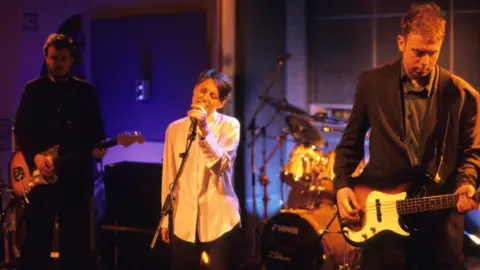 Getty Images Cocteau Twins performing on stage. Elizabeth Fraser is singing into a mic and there are band members on guitars and drums 