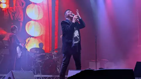 A man stands on stage and sings into a microphone. The stage has red and pink lighting and a band. The man has short dark hair and is wearing a black jacket.