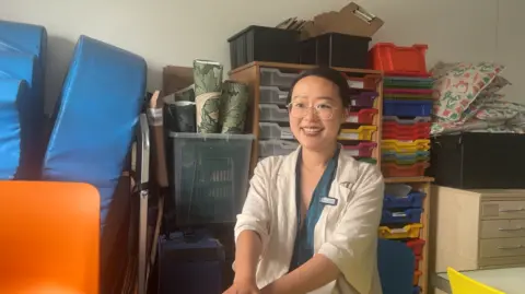 Jingyi Li has dark hair tied back. She is wearing round wire-rimmed glasses and a beige linen jacket. She is sat down and smiling in front of stacked, colourful plastic storage drawers.