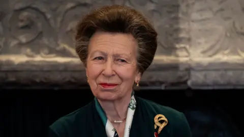 The Princess Royal, patron of The Butler Trust, attending the trust's annual awards at St James's Palace, London. She has brown hair and an emerald green velvet suit jacket, gold jewellery and red lipstick. 