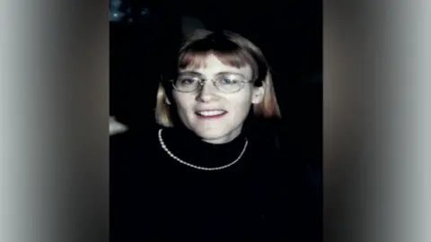 Family Handout Headshot of woman smiling at the camera. She is wearing a black turtleneck, glasses and a necklace.