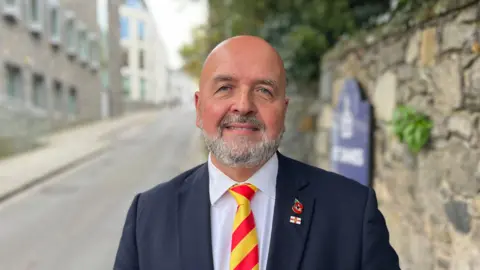 A bald man with a grey beard and moustache. He's wearing a blue suit and a white shirt with a bright red and yellow striped tie.