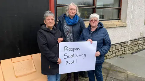 Three women outside a grey building, holding a campaign banner. Their sign says 'Reopen Scalloway pool'. 