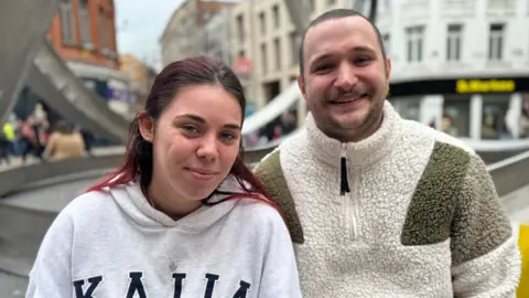 Caitlin and Samuel smiling at the camera. Caitlin has long red hair and is wearing a grey hoodie and silver necklace. Samuel has brown hair and is wearing a cream and green fleece.